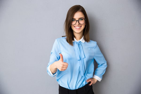 Happy businesswoman showing thumb up over gray background. Wearing in blue shirt and glasses. Looking at camera